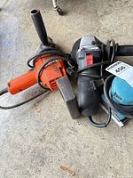 All three corded power tools lying on floor – drill, angle grinder with side handle, and sander with cords visible