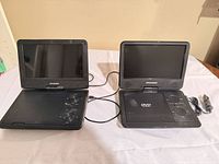 Front view of both Sylvania portable DVD players opened side by side with power adapter visible
