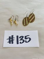 Both pairs of earrings laid flat: oval copper/brass weave pair and gold-tone seahorse pair, with lot number card visible