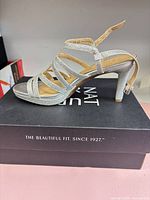 Side view of right sandal showing silver glitter finish, straps and heel on top of branded box