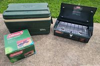 Coleman stove, Igloo cooler, boxed cookset grouped on pavement