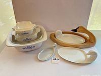 Group view of CorningWare casseroles stacked with lids, three ceramic swan dishes, and wooden swan tray