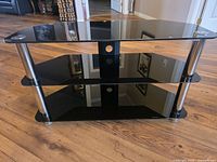 Front perspective of black glass three-tier TV stand on wood floor