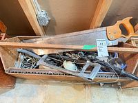 Overview of crate with hand saws, hacksaw, pipe wrench, electrical parts