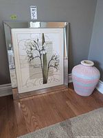 Mirrored framed floral print leaning against wall beside pastel textured ceramic vase on hardwood floor