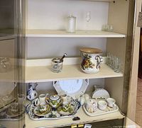 Cabinet shelves showing porcelain tea service on tray, charger plates, sugar bowl, ceramic pitcher and assorted clear glassware
