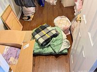 Stack with plaid blanket on green floral comforter, nearby bags and boxes of bedding
