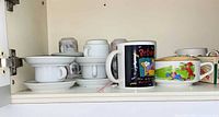 Stacked white demitasse cups and saucers, printed novelty mugs on cabinet shelf