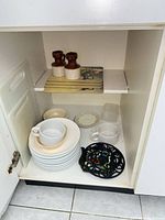 Cabinet view showing stacked plates, cups, saucers, bowl, cast iron trivet, placemats, creamers