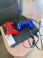 PS3 Slim console with red and blue controllers and power cord on top