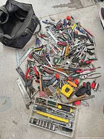 Overview of pile of assorted hand tools and black tool bag on floor
