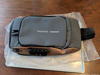 Front view of gray/brown sling bag showing FASHION logo and combination lock