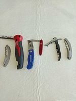 All six items laid out: Milwaukee utility knife, blue utility knife, Swiss Army penknife, three corkscrews