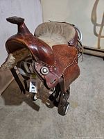 Front left view of saddle on stand showing floral tooling, suede seat, conchos
