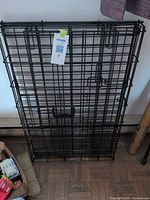 Folded black metal wire dog crate leaning against wall, MaxSold tag visible