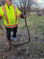 Man holding two scythes showing full length of wood handles and curved steel blades resting on ground