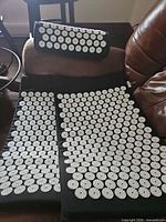 Two black acupressure mats with white spike discs and one matching pillow shown on sofa