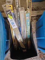 Multiple retail-packaged windshield wiper blades of various brands and sizes inside black fabric bag between blue bins