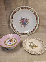 Front view of all three porcelain pieces: large floral dinner plate, pink pedestal compote, small scenic plate