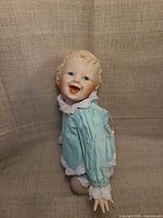 Front view of doll seated, showing porcelain head, hands, blue dress, facial details