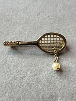 Full view of gold-tone tennis racquet brooch with dangling faux pearl