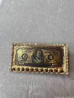 Front view of gold-tone brooch with rhinestone border and money design