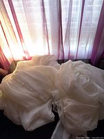 Piled white sheer curtain panels on dark surface with backlit window showing translucence