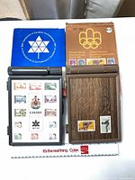 Both stamp sets with open Centennial case showing mounted stamps, closed Olympic case, and respective cardboard boxes