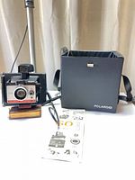 Front view of Polaroid Land Camera Colorpack 80 on stand beside carrying case and manual