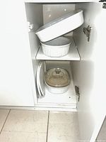 Cabinet view showing two round casseroles on upper shelf and square casserole with glass lid, plastic lids and platter on lower shelf