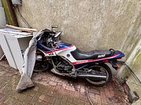 Whole motorcycle against wall with partial cover and nearby debris