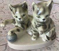 front view of glazed ceramic figurine showing two grey tabby cats with blue-grey yarn ball on cream oval base