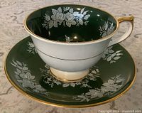 Teacup on saucer, exterior view showing dark green glaze, white floral pattern, gold trim