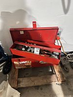 Red steel toolbox on pallet, lid open showing assorted tools