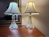 Two matching crystal base table lamps with shades plugged in on wooden surface