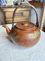 Front view showing hammered copper body, floral gilt motifs, lid in place and upright handle