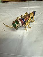 Left side view of cloisonné grasshopper ornament showing multicolour enamel sections and gold-tone legs