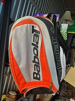 Front and side view of orange, white and black Babolat backpack hanging, showing overall design