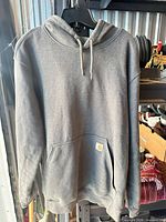 Front view of grey Carhartt pullover hoodie on hanger showing hood, drawstrings, kangaroo pocket, logo patch