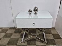 Front view of side table showing glass top, drawer, and stainless steel cross legs