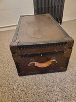 Closed trunk angled view showing top, leather side handle, metal corner brackets