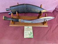 Kukri knife on wooden stand, sheath above knife, certificate in front