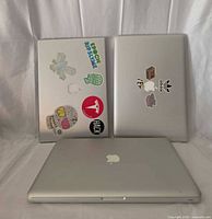 Three closed MacBook Pro laptops, two with sticker-covered lids, showing Apple logos