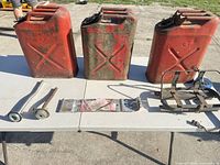 Three red/green steel jerry cans, two spouts, strap and carrier on table