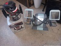 Router on box next to bench grinder with attached cords, eye shields and tool rests visible