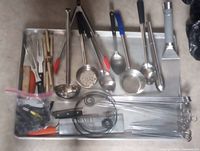 Full view of tray containing assorted stainless steel ladles, spoons, tongs, grill scraper, carving forks, skewer sets, peeler, knife, and bagged accessories