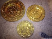 Three brass decorative wall plates with embossed scenes displayed on carpet