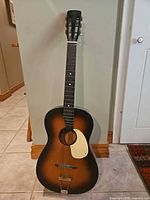 Full front view of guitar leaning against wall showing sunburst finish, pickguard and tailpiece