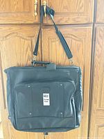 Full view of black garment bag hanging, showing front pockets and shoulder strap
