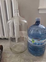 Side view of clear glass carboy beside 15 L plastic bottle for scale